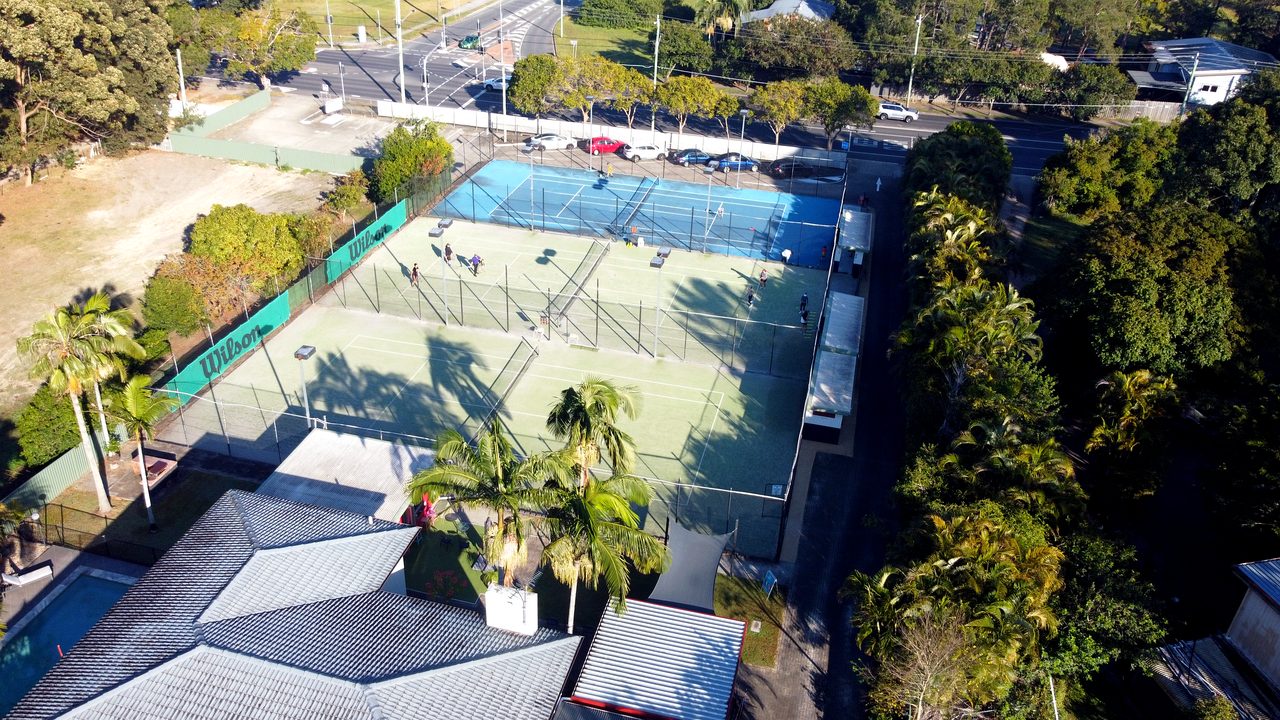 Aerial view of Logan Tennis Club — two synthetic grass courts, the blue hard court and the clubhouse
