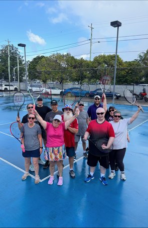 About a dozen adults on the blue hard court holding their racquets up and smiling, with the car park and floodlights behind