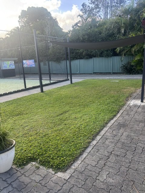 A grassed area with a shade sail beside the fenced synthetic grass court