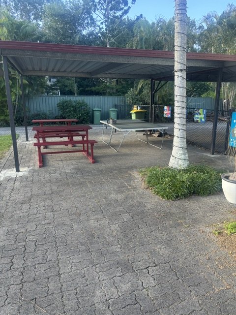 A covered pergola with a red picnic table and a table-tennis table on pavers beside the courts