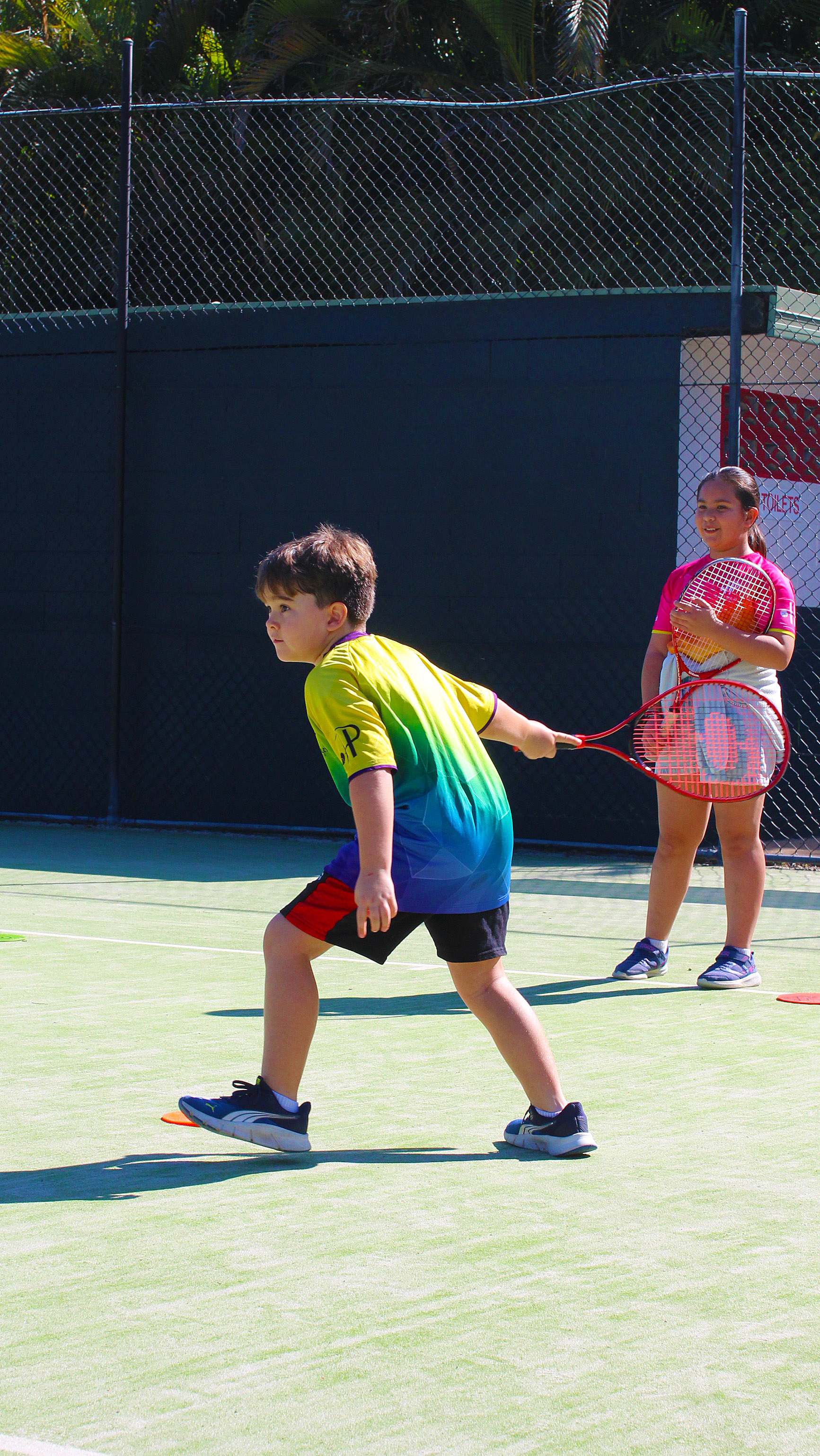 A junior in a rainbow club shirt follows through with a red racquet while another player waits behind