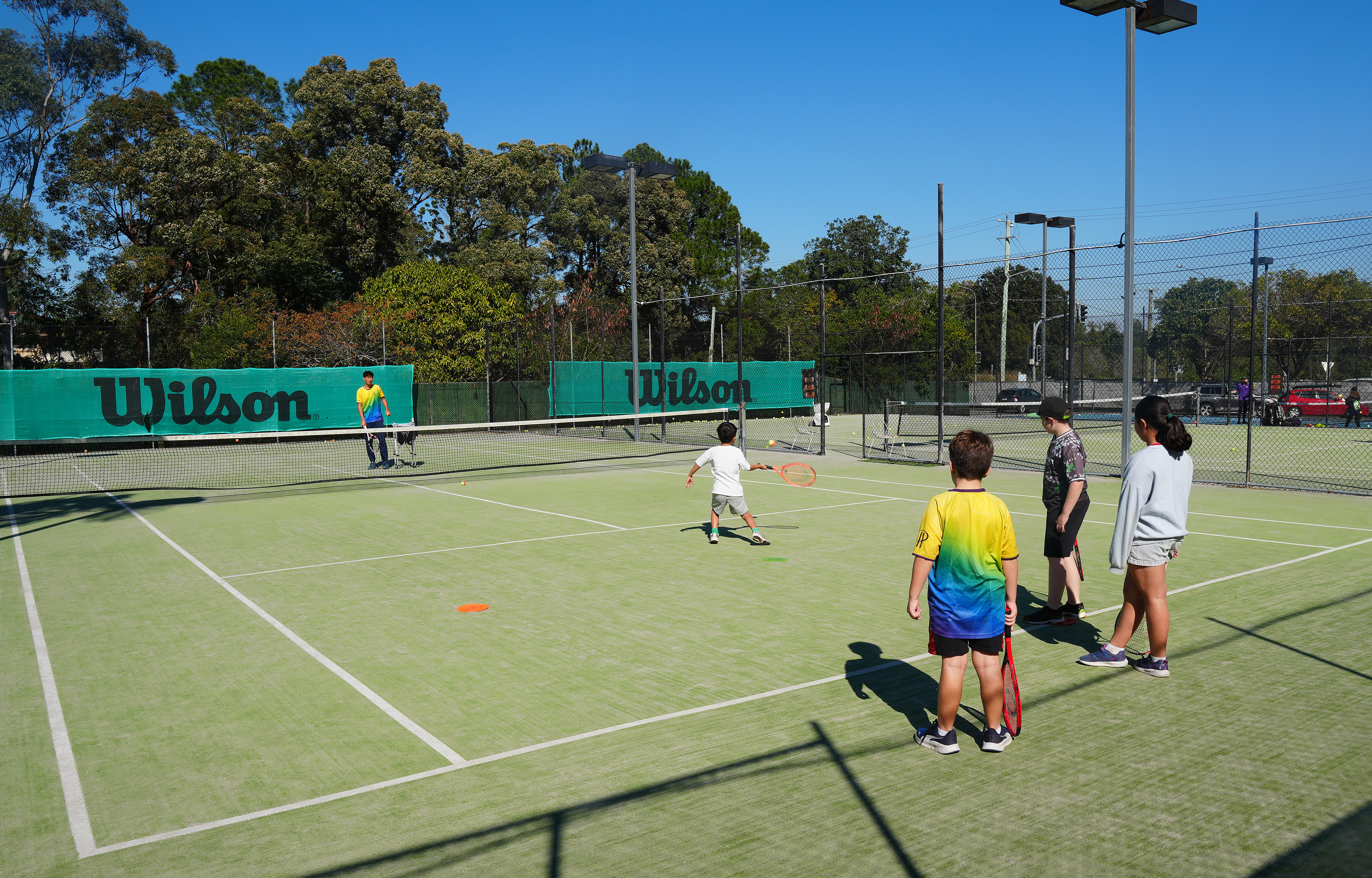 A junior reaches for a ball near the net while three other players wait on the synthetic grass court
