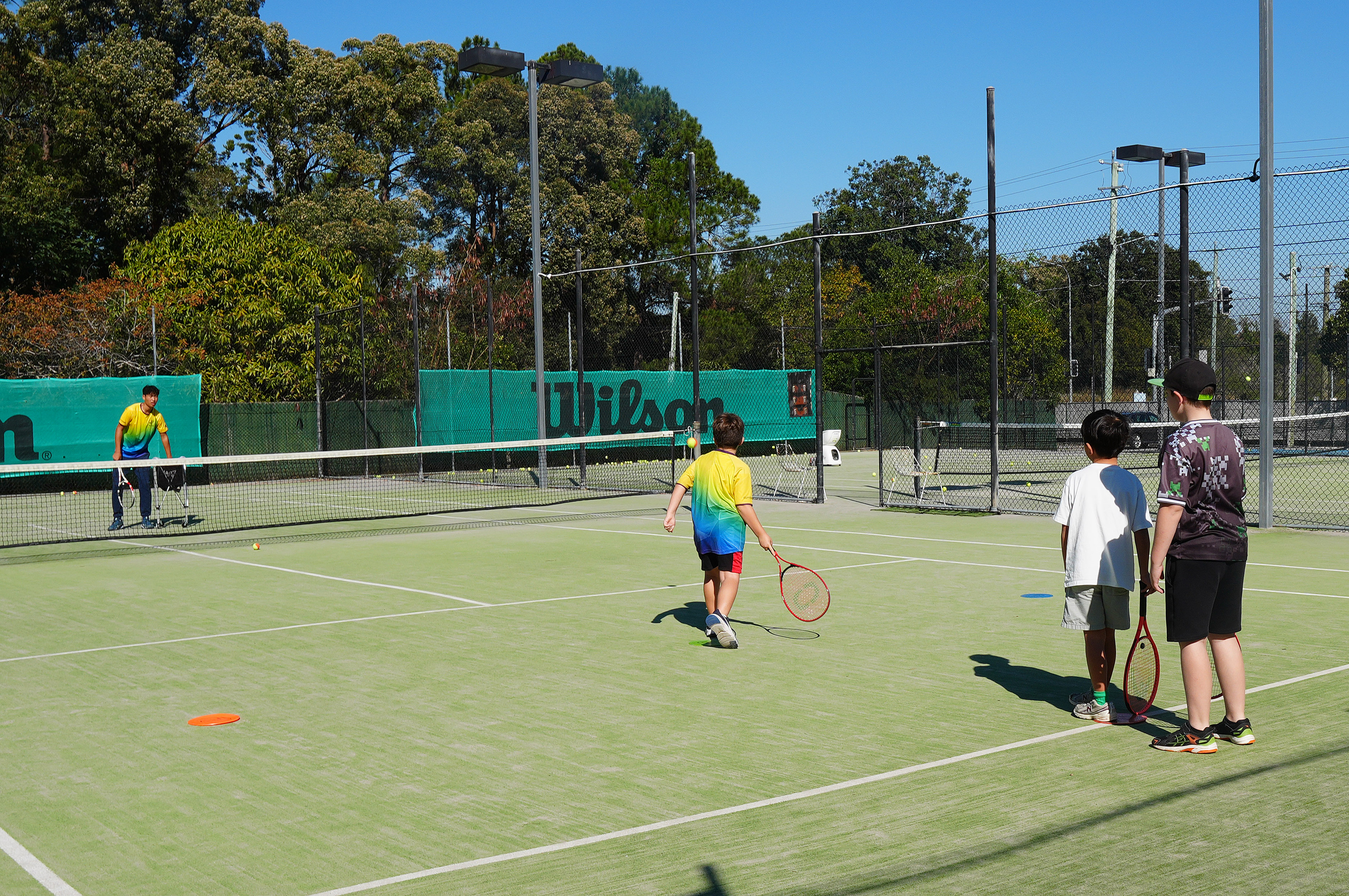 A junior in a rainbow club shirt steps towards the net as two other players wait with their racquets