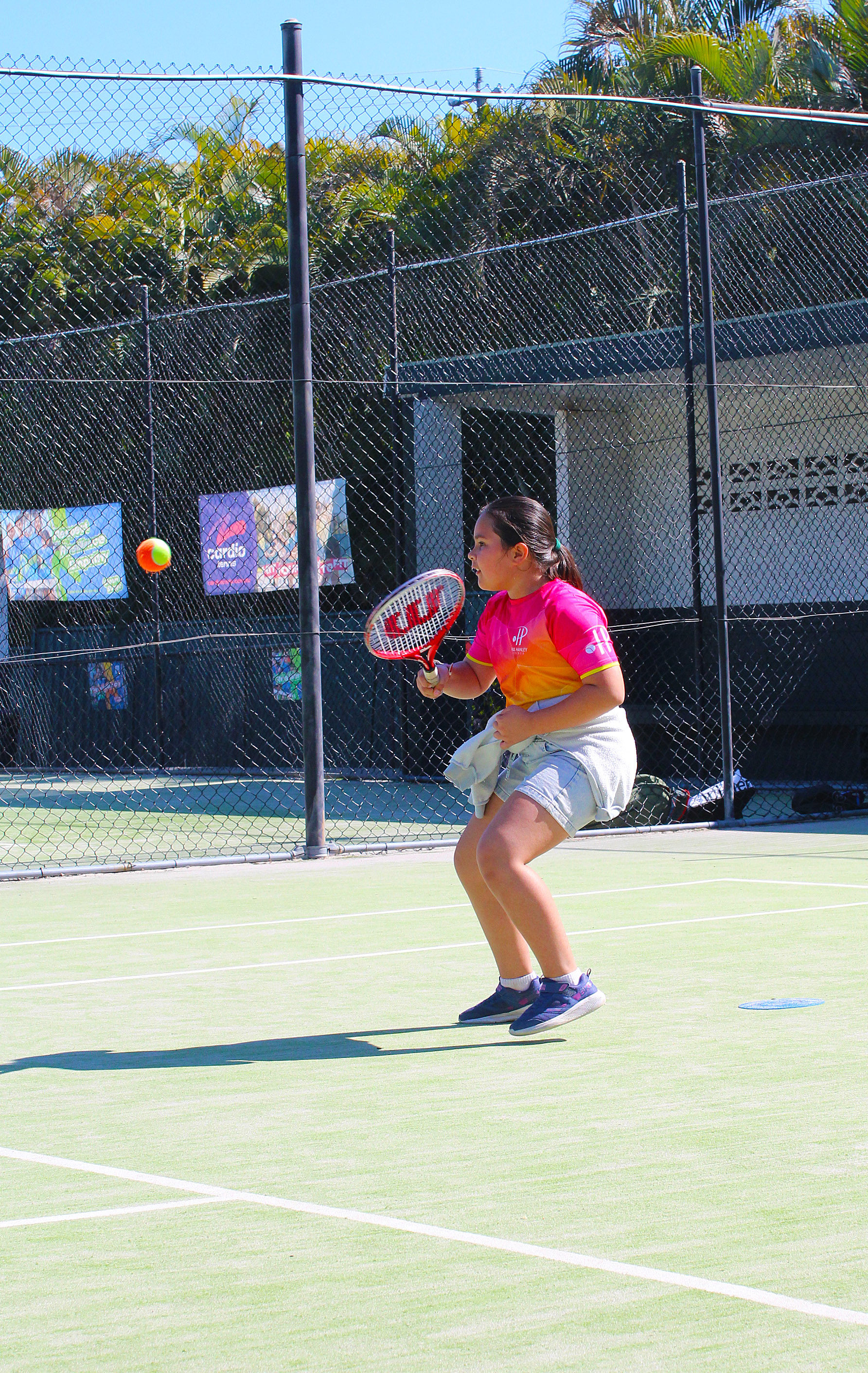 A junior in a pink and orange club shirt prepares to return an orange tennis ball