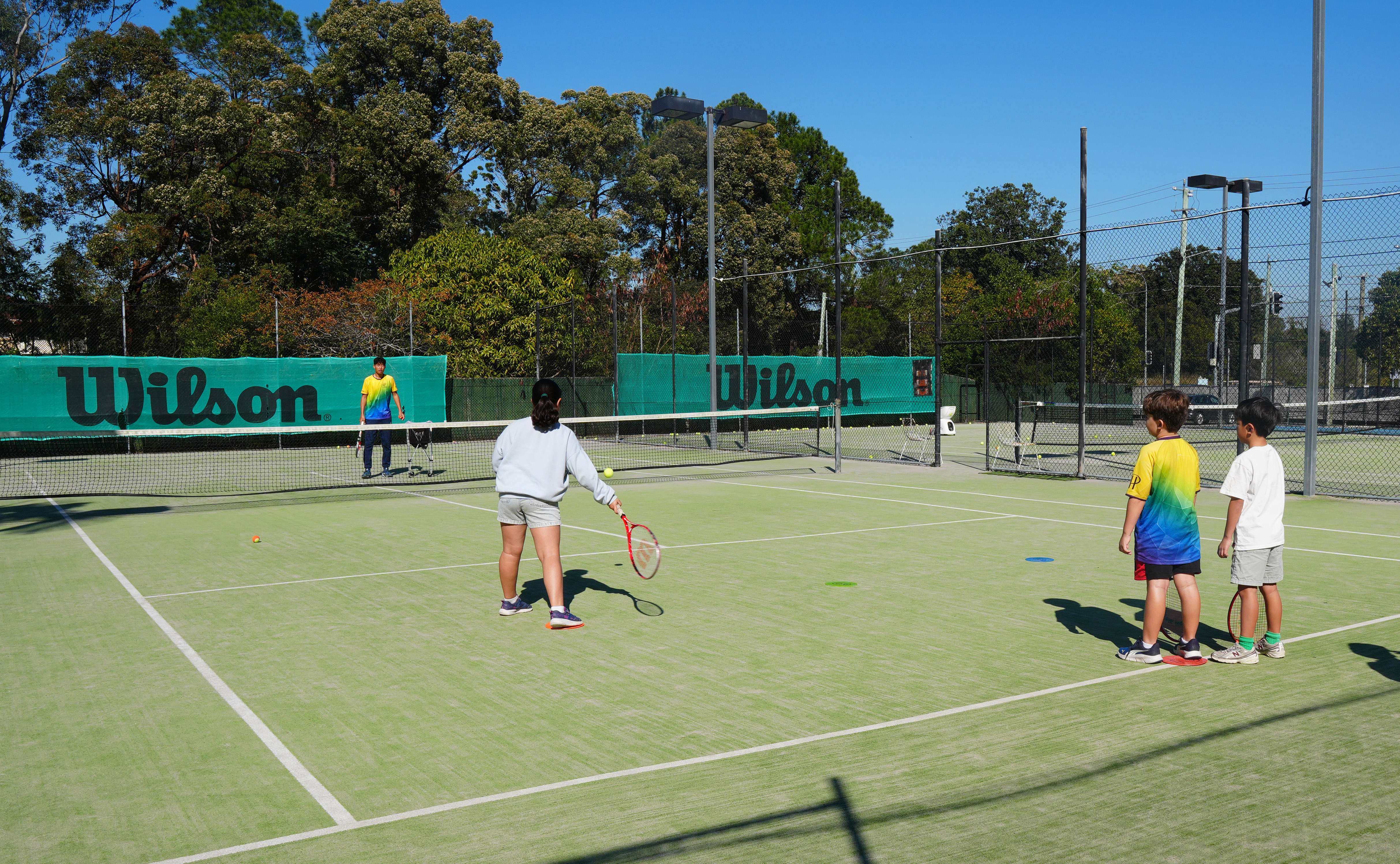 A junior in a grey top reaches towards a ball near the net while two other juniors wait to take their turn