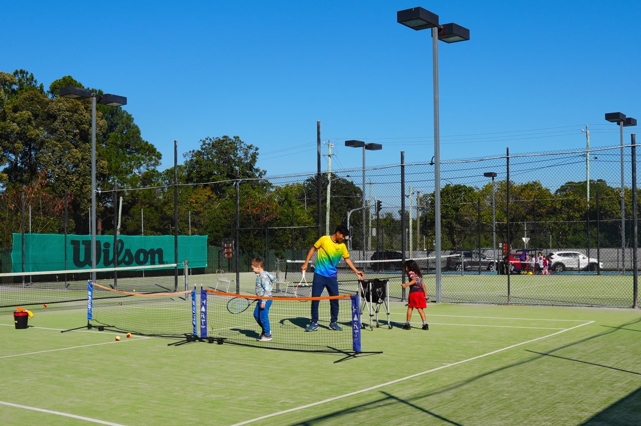 A coach in a club shirt feeds balls over an orange mini net to two young players on the synthetic grass court