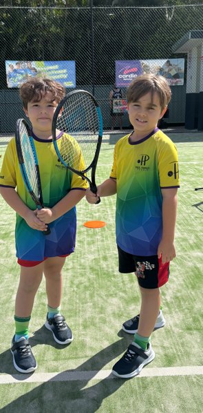 Two boys in matching rainbow club shirts on the synthetic grass court, one holding a racquet