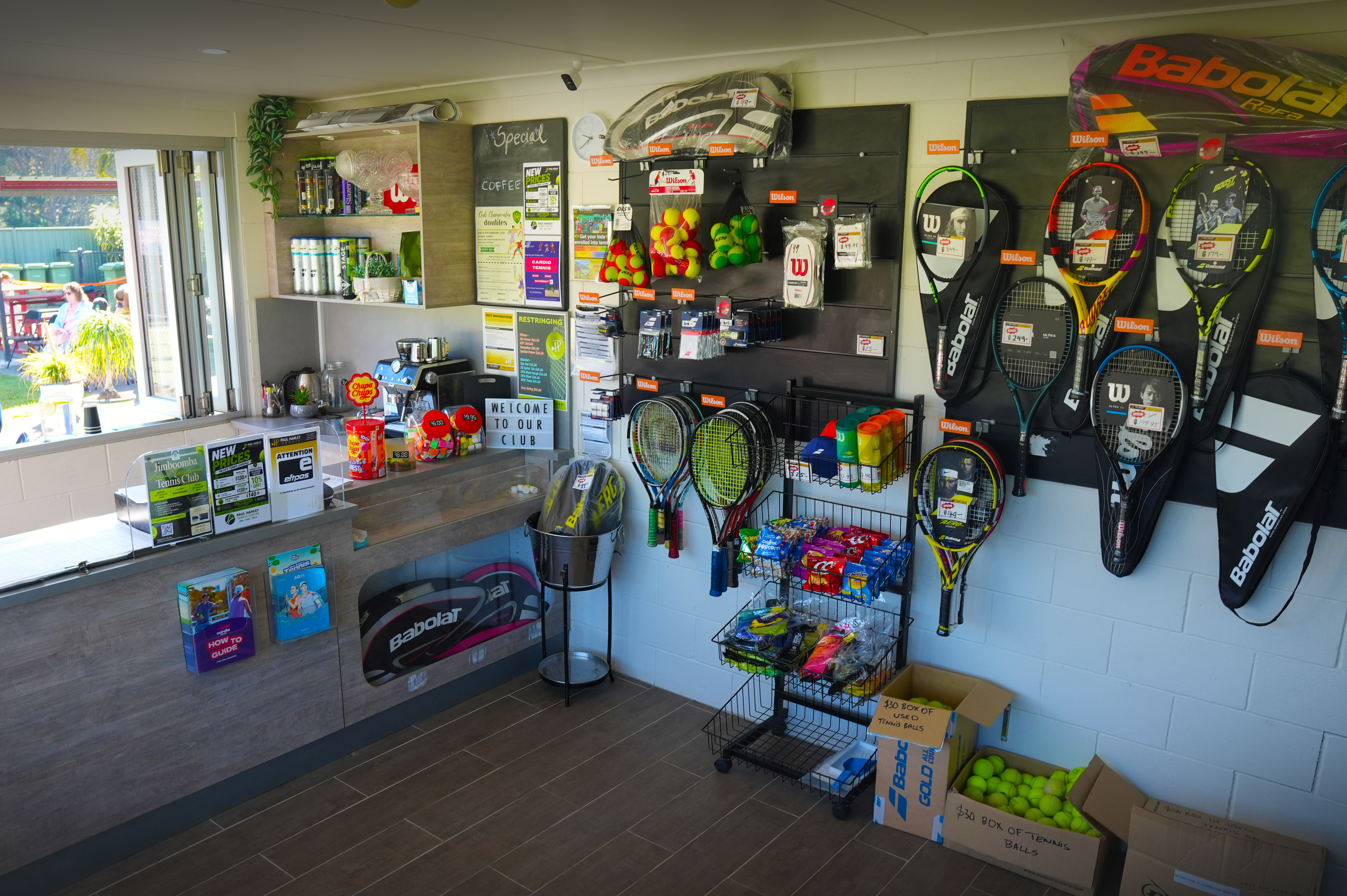 Racquets, tennis balls, bags and accessories displayed beside a counter and coffee machine in the pro shop