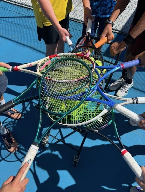 Six racquets fanned over a basket of tennis balls on the blue hard court, hands holding the grips